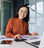Portrait of young Asian successful financier working on paperwork with calculator inside office, businesswoman smiling and looking at camera, filling forms and verifying financial reports.