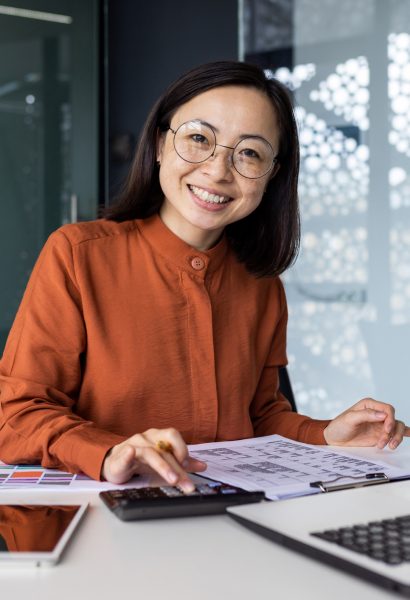 Portrait of young Asian successful financier working on paperwork with calculator inside office, businesswoman smiling and looking at camera, filling forms and verifying financial reports.