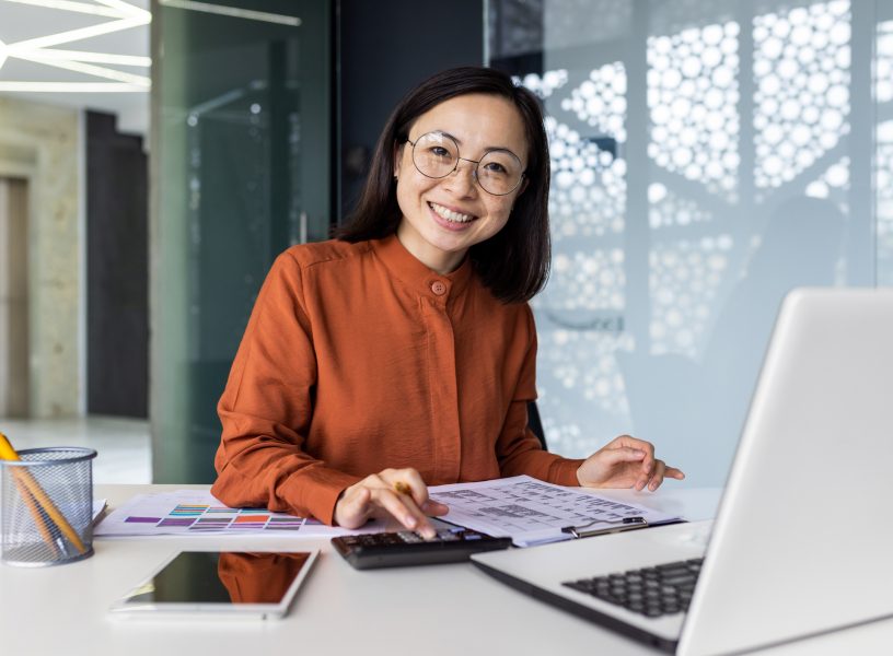 Portrait of young Asian successful financier working on paperwork with calculator inside office, businesswoman smiling and looking at camera, filling forms and verifying financial reports Portrait of young Asian successful financier working on paperwork with calculator inside office, businesswoman smiling and looking at camera, filling forms and verifying financial reports.