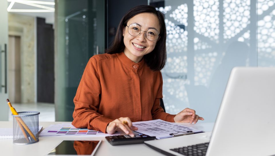 Portrait of young Asian successful financier working on paperwork with calculator inside office, businesswoman smiling and looking at camera, filling forms and verifying financial reports.