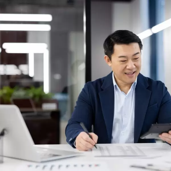 Smiling chinese accountant summarising expenses and income while using calculator and writing down indicators. Attentive economist filling document with tables during busy day in office.