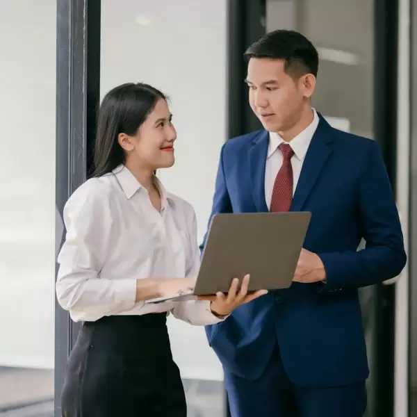 A woman and a man are standing in front of a window, with the woman holding a laptop. They are both dressed in business attire, and the man is wearing a tie. Scene is professional and focused
