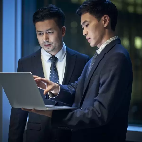 two asian corporate executives standing by the window in office discussing business using laptop computer at night